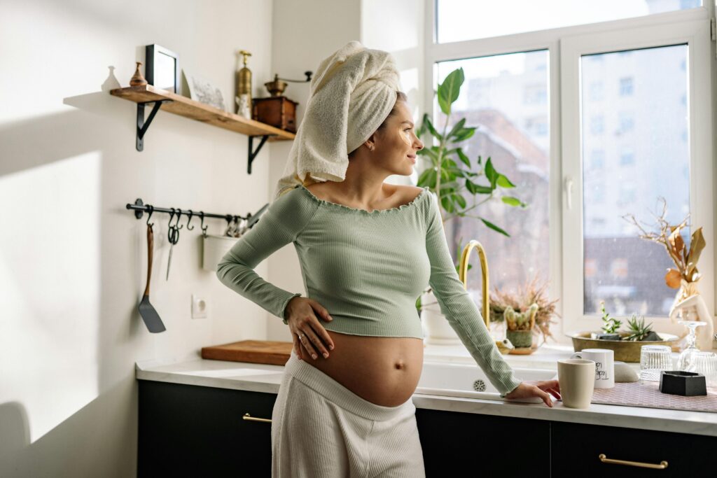 Pregnant woman standing in the kitchen, looking happy. Foods to avoid for you and your baby well-being.