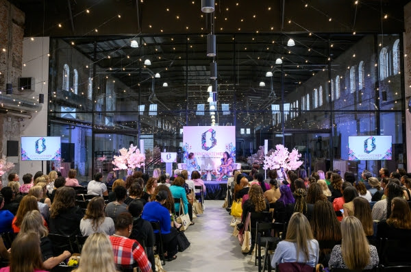 A wide shot shows a packed audience at The Baltimore Banner's InnovateHER event, attentively watching a panel discussion on a brightly lit stage. The stage backdrop features a stylized "B" logo with floral accents and the vertical text "BOSSBABE," flanked by large screens displaying the same branding. Four women are seated at a table on stage, engaged in conversation, positioned in front of the backdrop and artificial cherry blossom trees. The audience, primarily women, fills rows of black chairs in the large, industrial-chic venue with exposed brick, high ceilings, and string lights. The atmosphere is focused and engaged, indicative of a conference celebrating and empowering women in innovation.